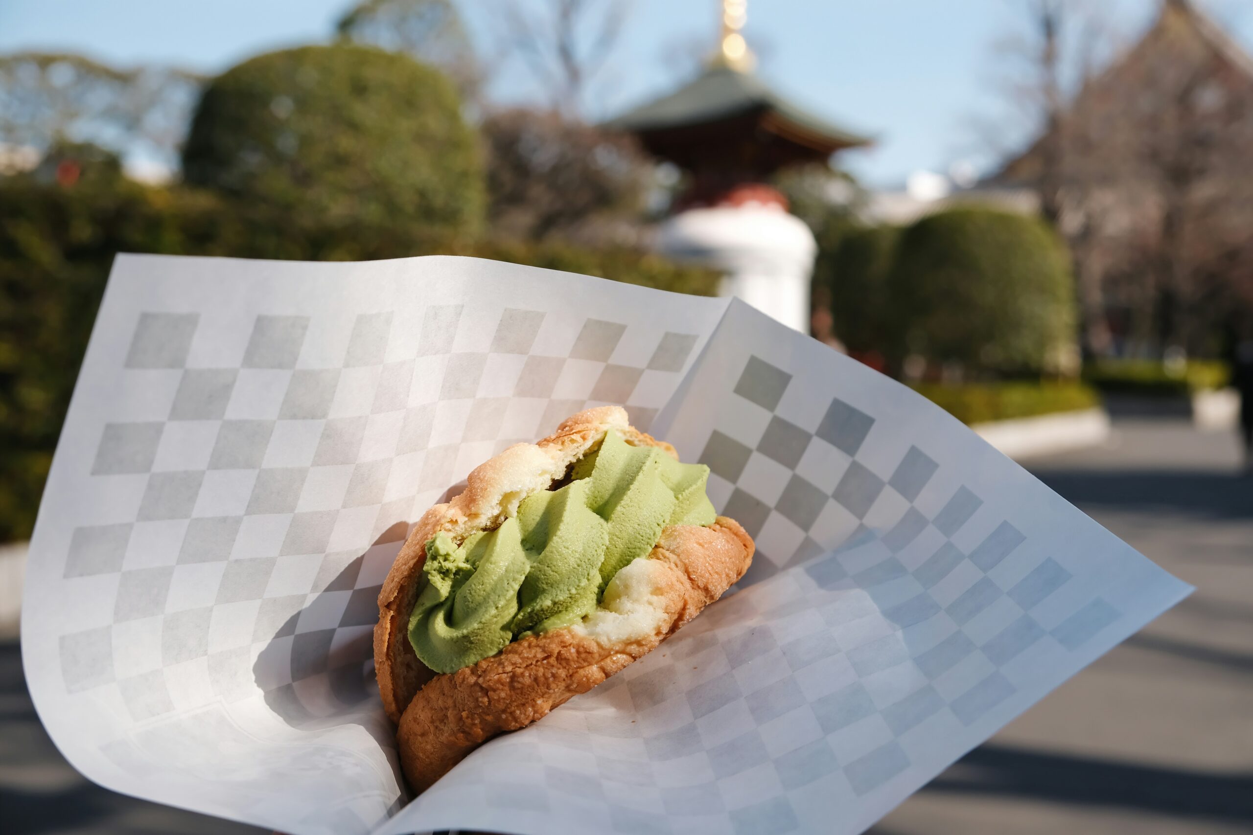 Matcha soft serve in a cream puff held in checkered paper, with a temple in the background — street food to eat while walking